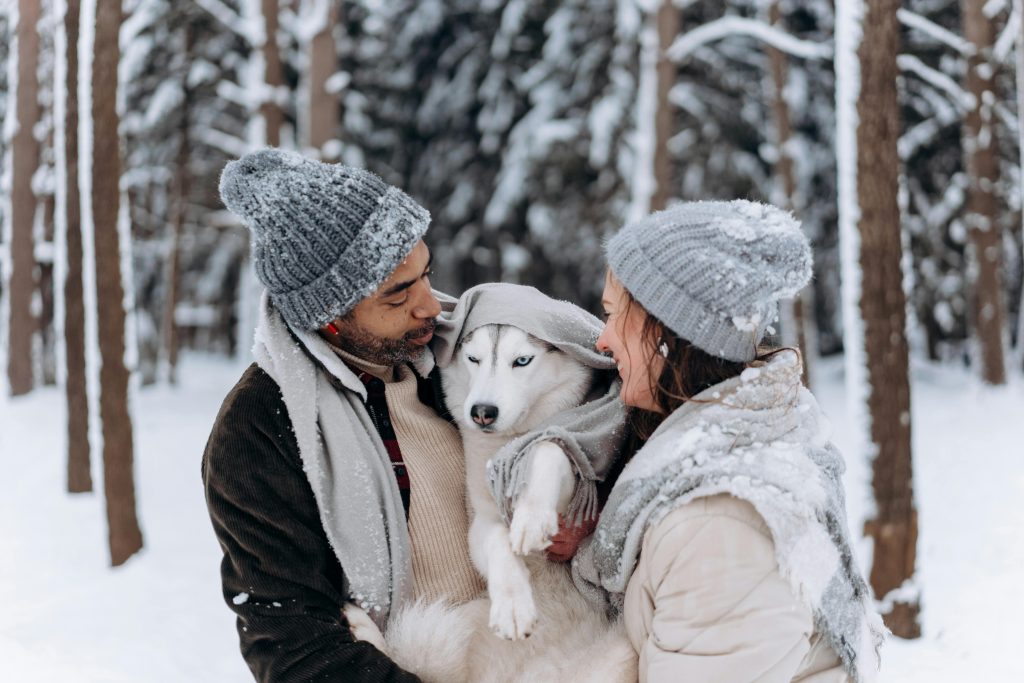 A couple in winter clothing embraces their husky dog in a snowy forest.