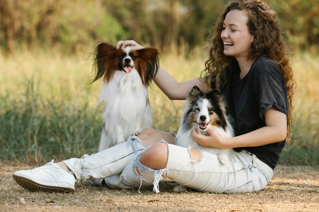 A joyful woman playing with a Papillon and Sheltie dog outdoors, smiling warmly in the sunshine.