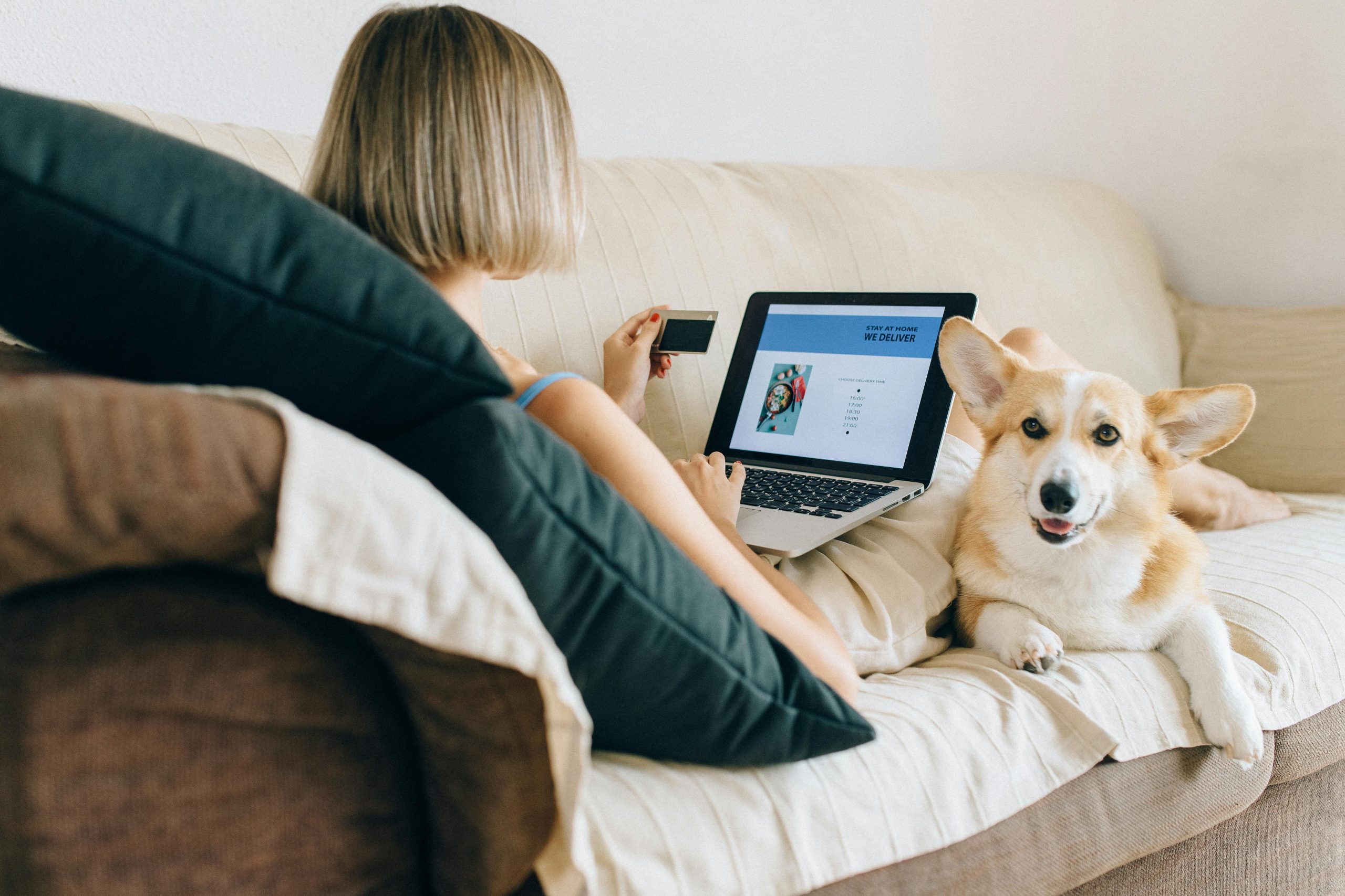 A woman using a laptop for online shopping with her corgi dog beside her on the couch.